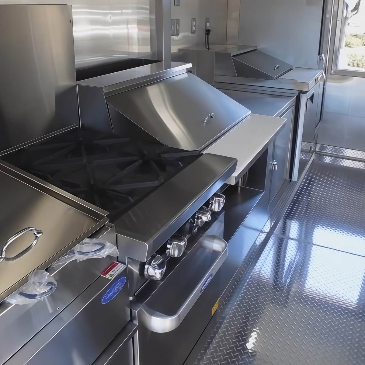 Interior view of a commercial food trailer featuring a four-burner cooking range, large commercial stove, dual refrigeration units, and stainless steel kitchen equipment for mobile catering operations.
